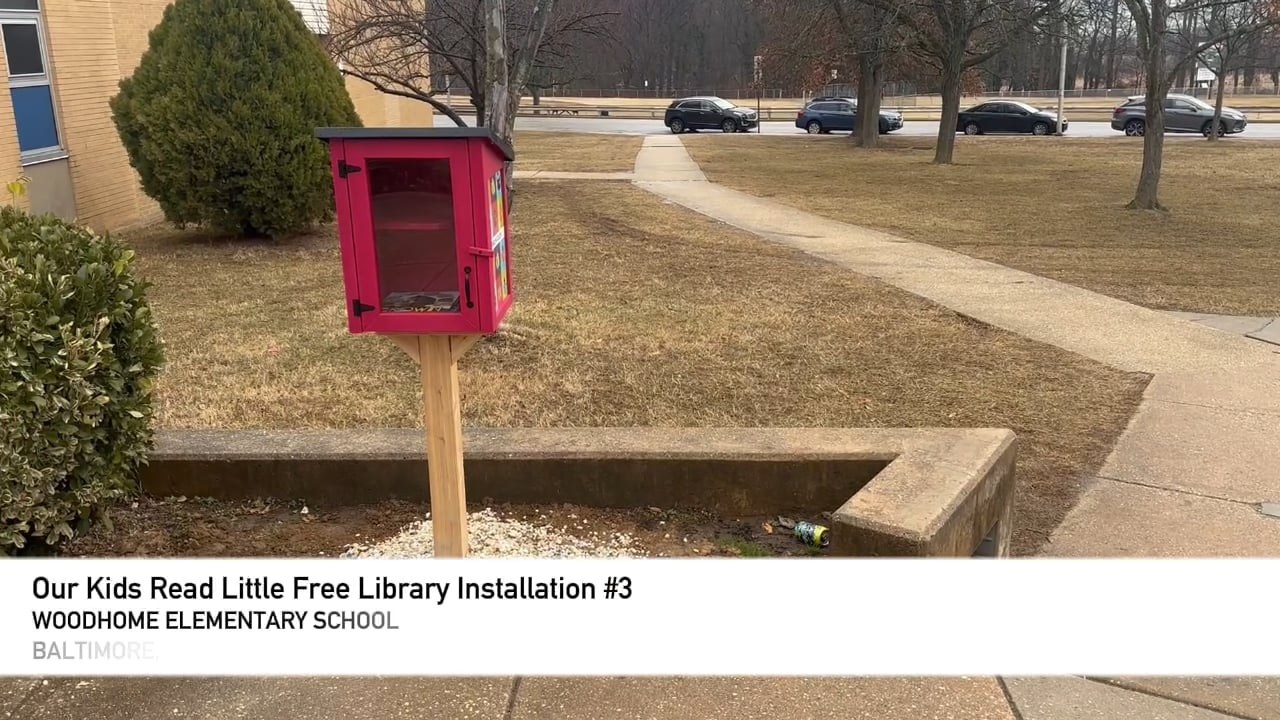 A red Our Kids Read Little Free Library installed on the lawn at Woodhome Elementary School in Baltimore